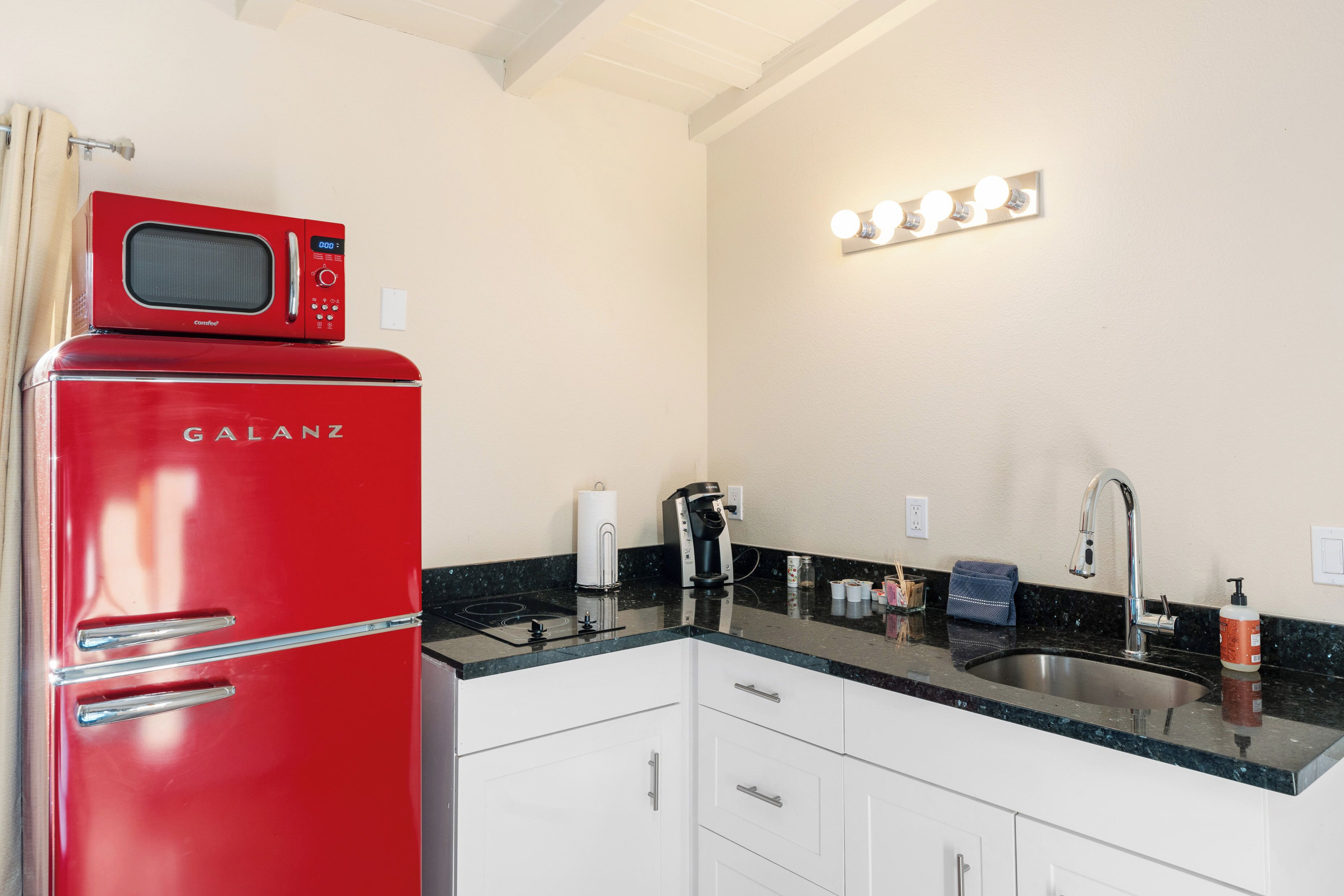 A kitchen with a red Galanz refrigerator, a black countertop, white cabinets, a sink, and a lighted mirror above the countertop.