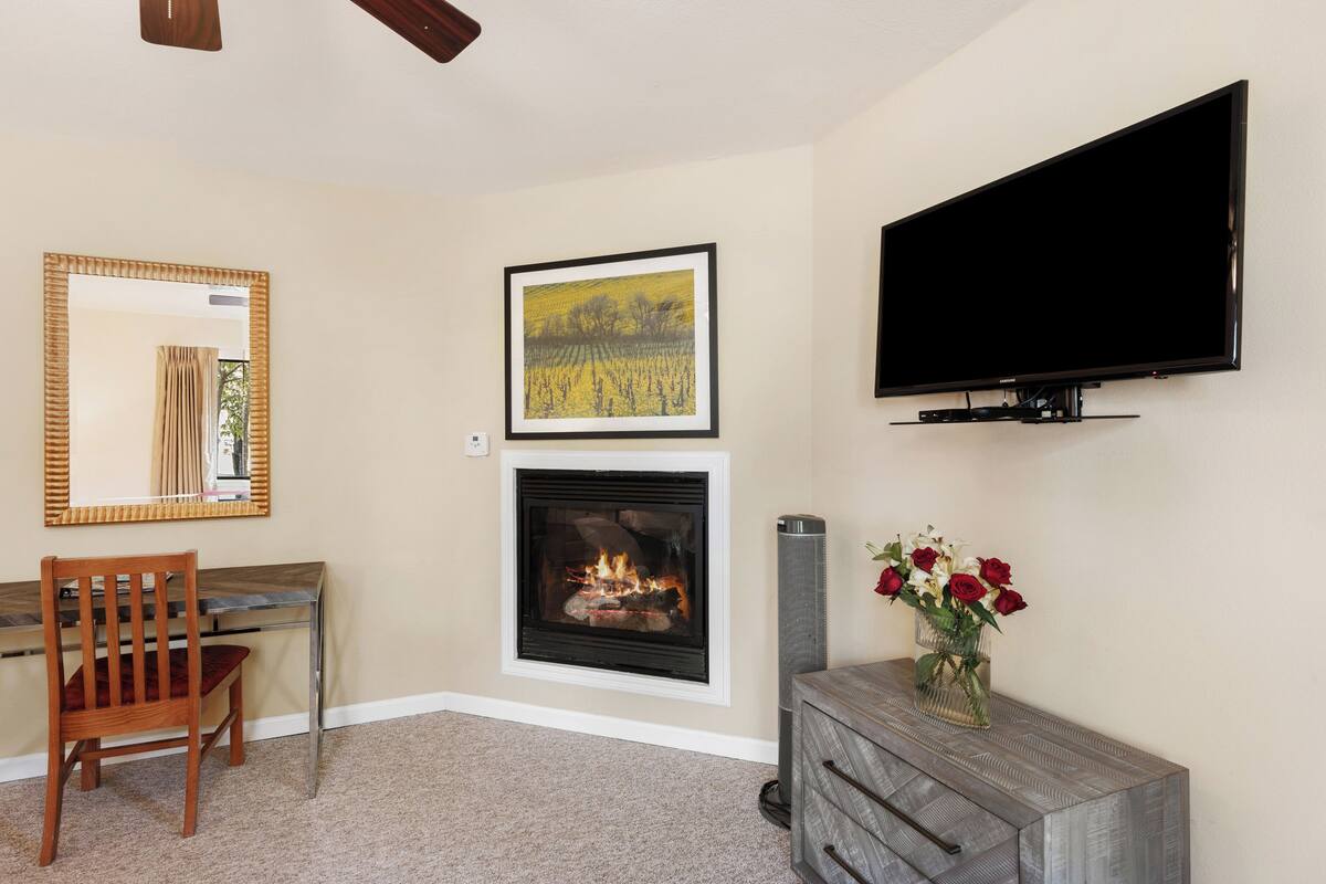 A living room with a fireplace, a television mounted on the wall, a framed picture above the fireplace, and a vase with flowers on a side table.
