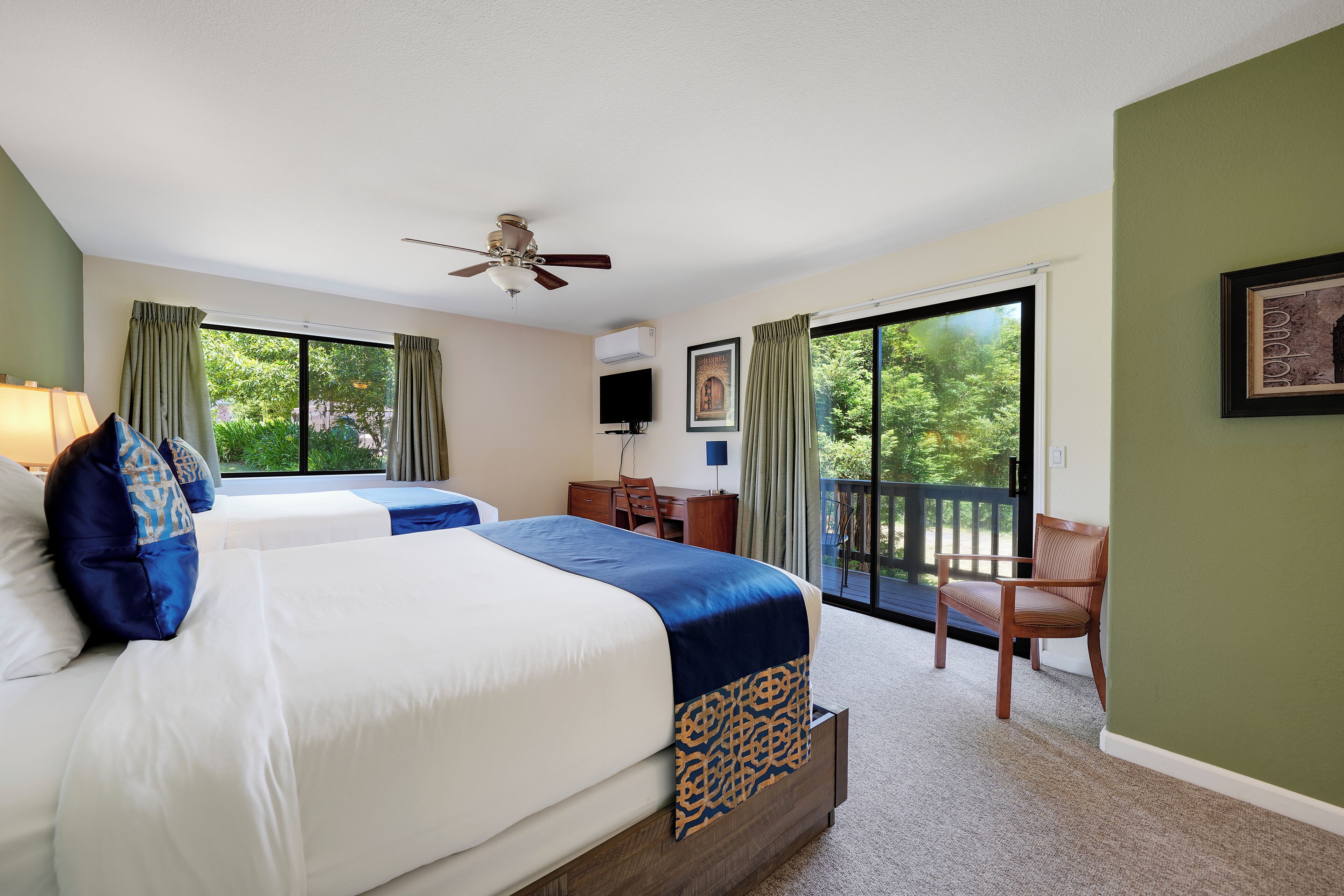 A hotel room with two beds, a ceiling fan, a TV, and a balcony with a chair.