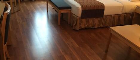 A hotel room with a wooden floor, a bed with a brown bedspread, a bench, a bedside table, and a lamp.