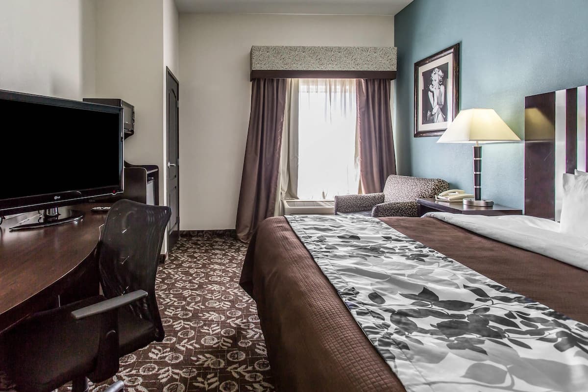 A hotel room with a bed, a desk with a TV, a chair, a lamp, and a window with curtains.