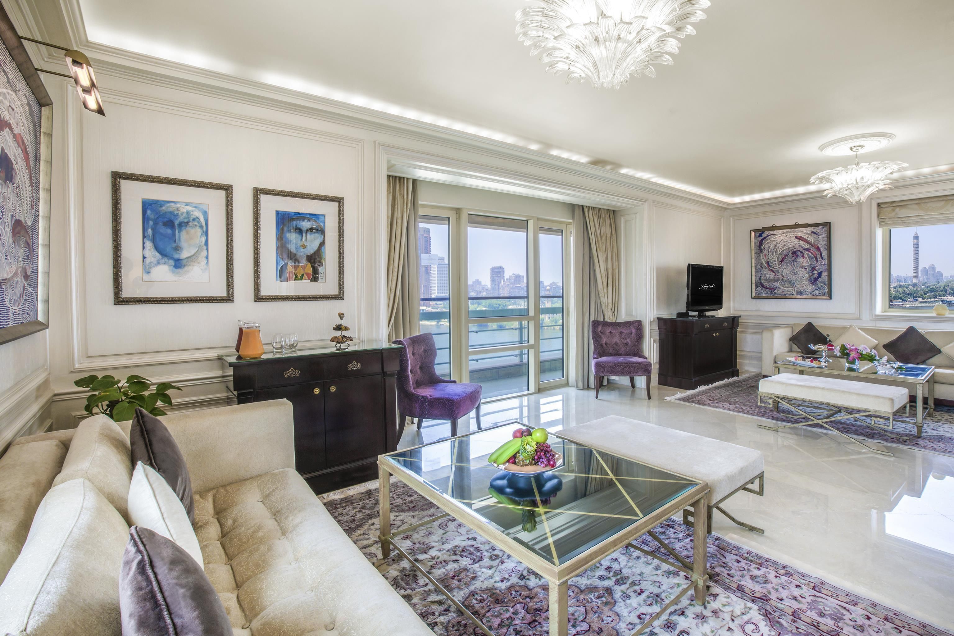 Un salón amplio con sofá, sillones, una mesa de centro y televisión. Hay ventanas grandes con vistas a un paisaje urbano.