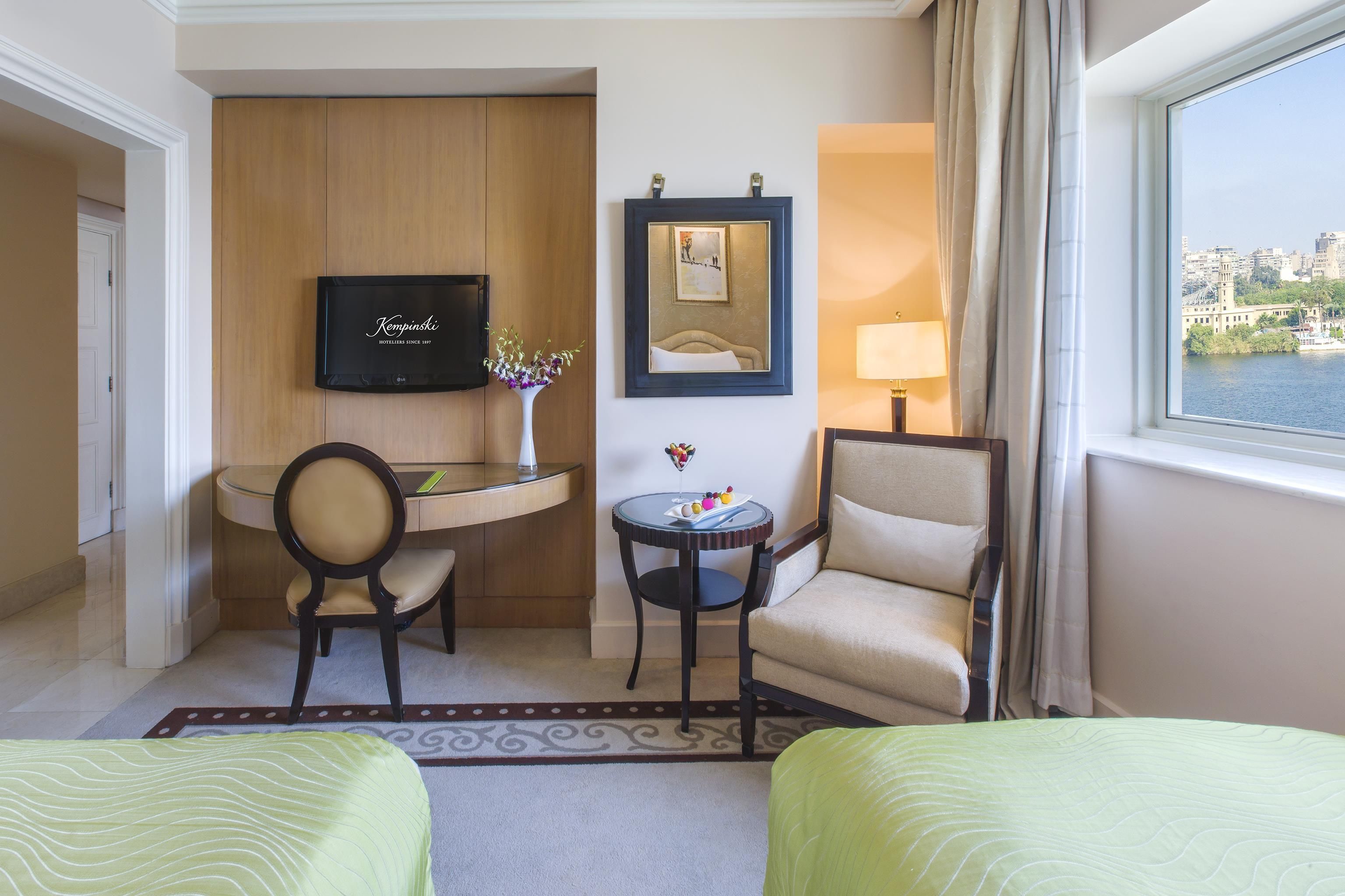 A hotel room with two beds, a desk, a chair, a TV, a mirror, and a window with a view of the city.