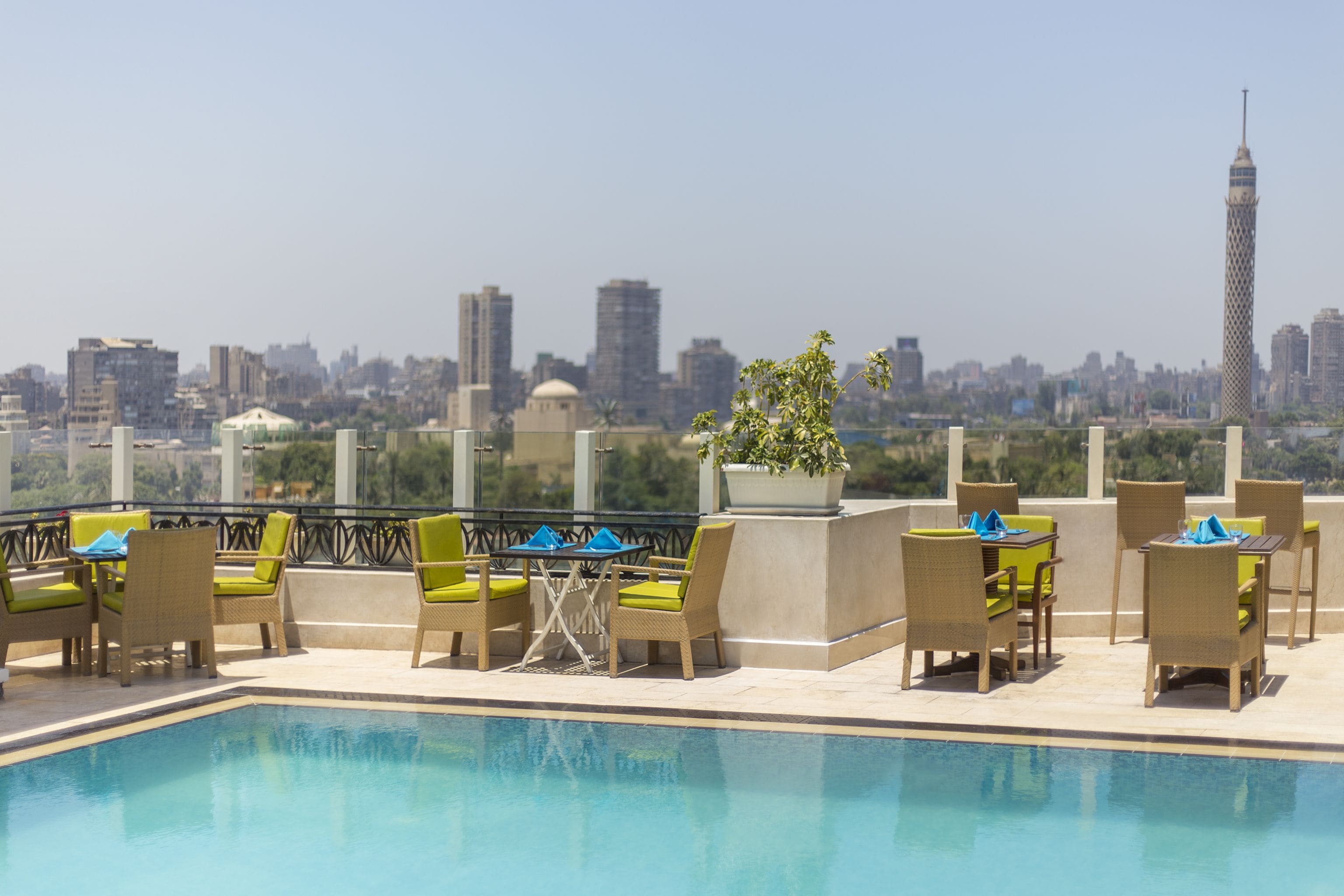 A rooftop pool area with lounge chairs and tables, a view of a city skyline, and a prominent tower in the distance.