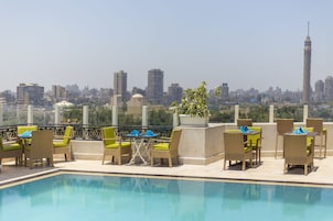 A rooftop pool area with lounge chairs and tables, a view of a city skyline, and a prominent tower in the distance.