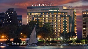 A modern hotel with multiple floors, a sailboat in the foreground, and a cityscape in the background. - Kempinski Nile Hotel Cairo (Cairo)