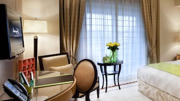 A hotel room with a bed, a desk with a lamp, a chair, a TV, a vase with flowers, and a window with curtains.