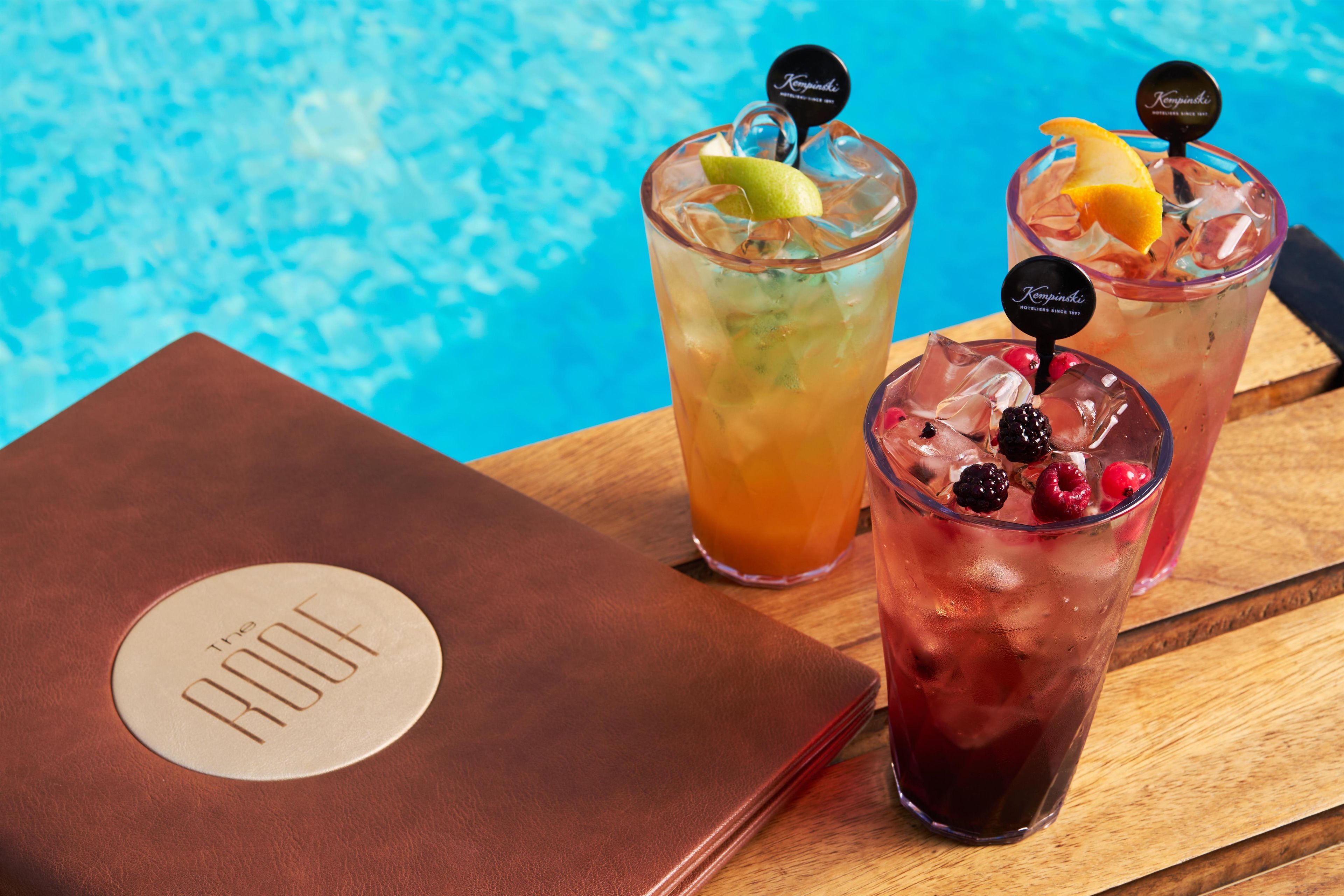 Three iced beverages with fruit garnishes and a leather-bound menu on a wooden table by a pool.
