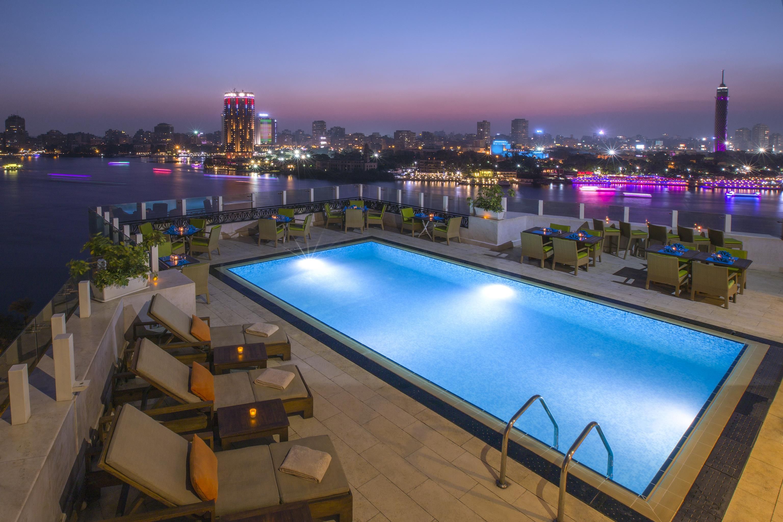 A rooftop pool area with lounge chairs and tables, overlooking a cityscape at dusk.