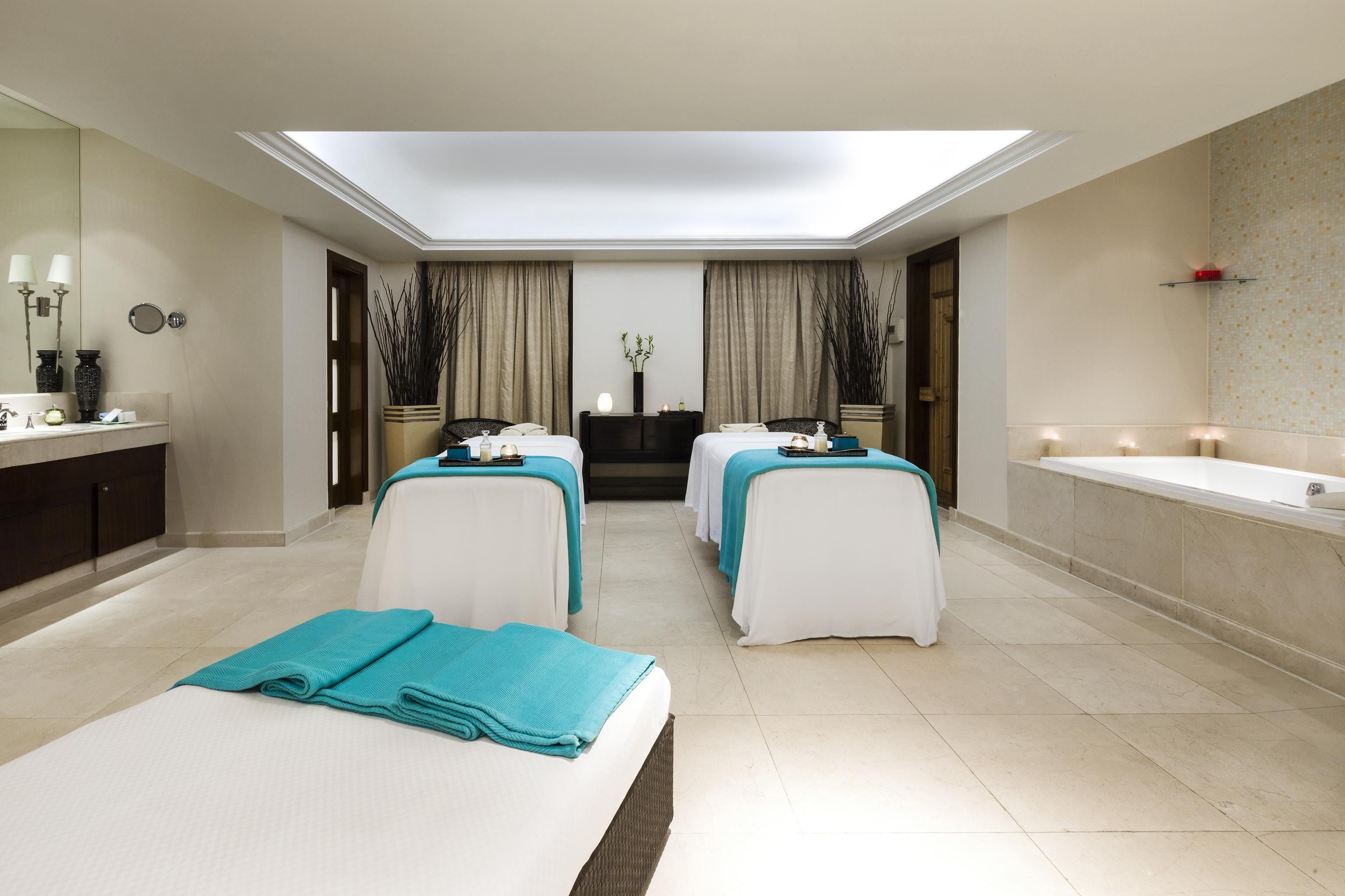 A modern spa room with two massage tables, a large mirror, and a bathtub.