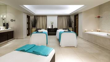 A modern spa room with two massage tables, a large mirror, and a bathtub.