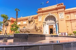 A large stone sphinx statue in front of a building with classical architecture, palm trees, and flags. - Kempinski Nile Hotel Cairo (Cairo)