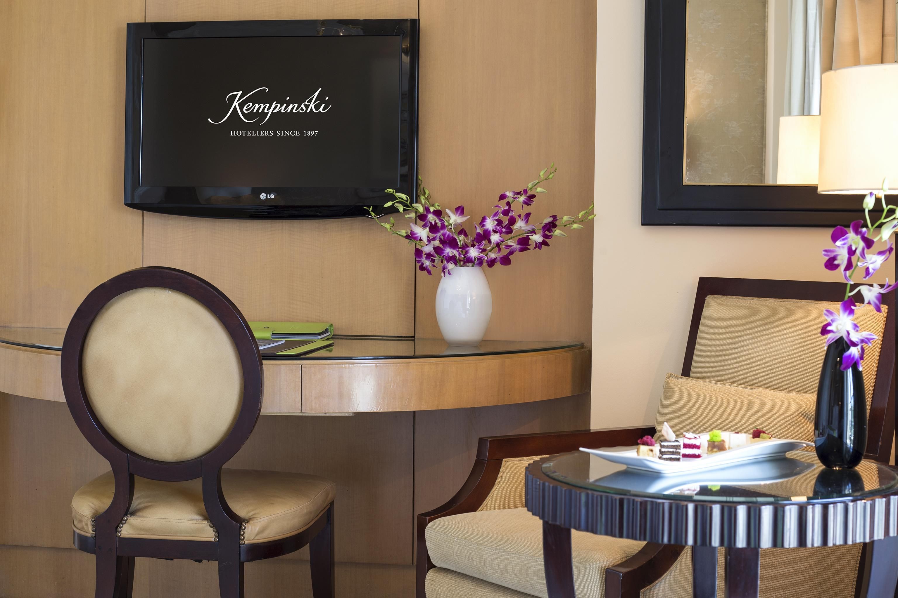 A hotel room with a television displaying 'Kempinski HOTELIERS SINCE 1897', a vase with purple flowers, a round table with a tray of desserts, and a chair with a cushion.