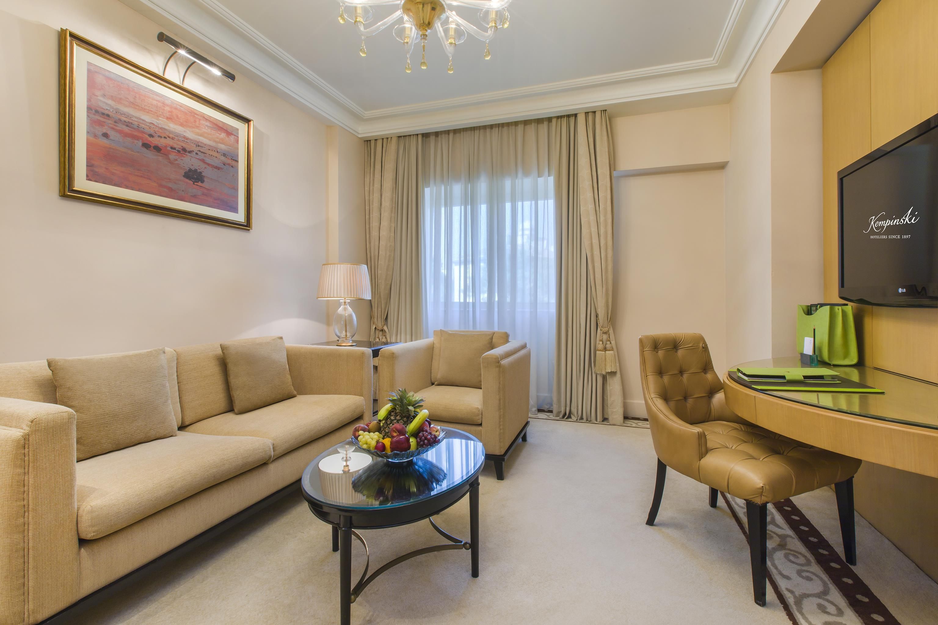 Une chambre d’hôtel avec un canapé, une table basse avec des fruits, un fauteuil, un bureau avec un écran et une grande fenêtre avec des rideaux.