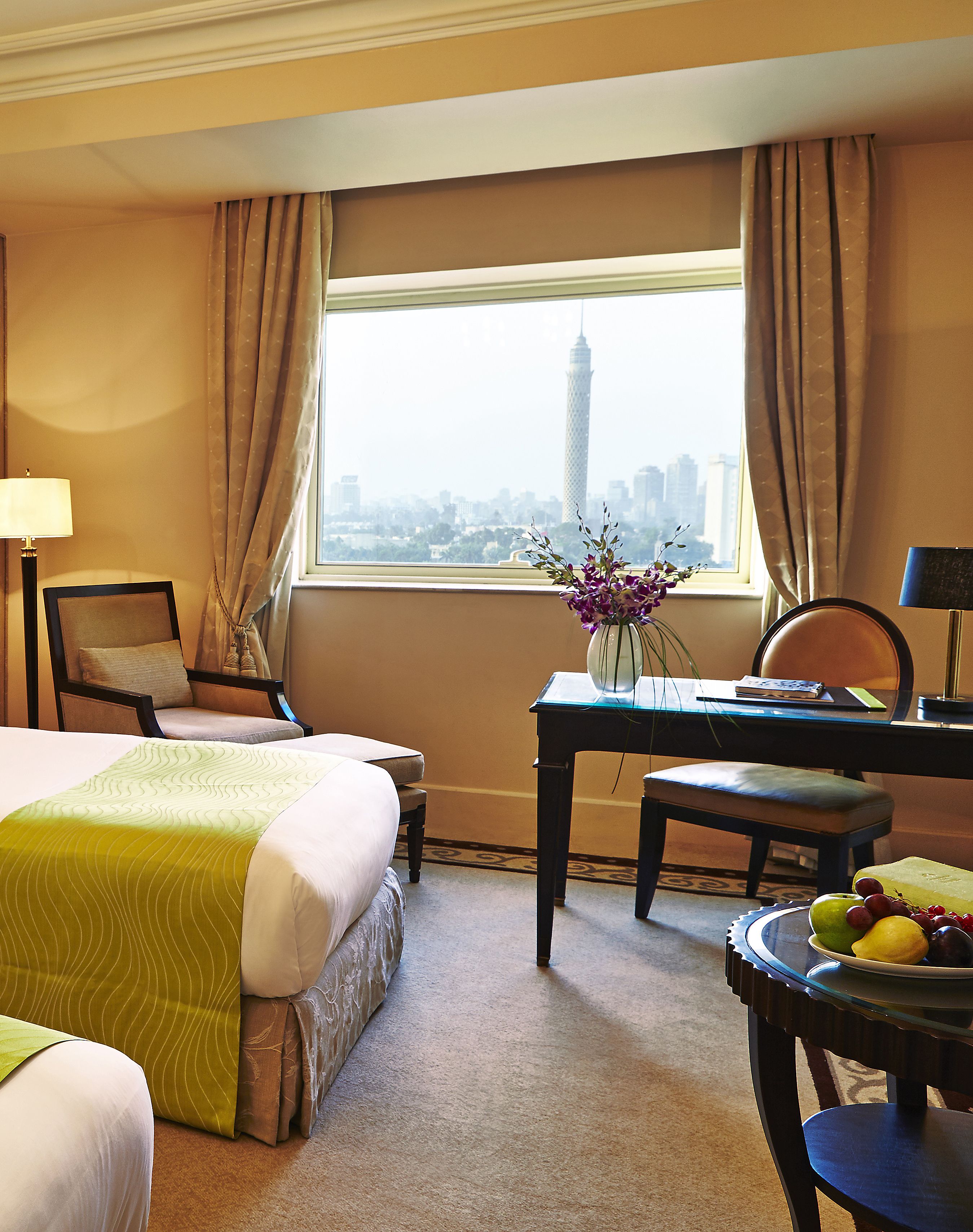A hotel room with a bed, a desk, a chair, a table with fruit, and a view of a cityscape.