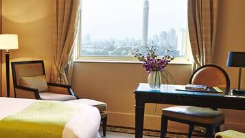 A hotel room with a bed, a desk, a chair, a table with fruit, and a view of a cityscape.