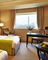 A hotel room with a bed, a desk, a chair, a table with fruit, and a view of a cityscape.