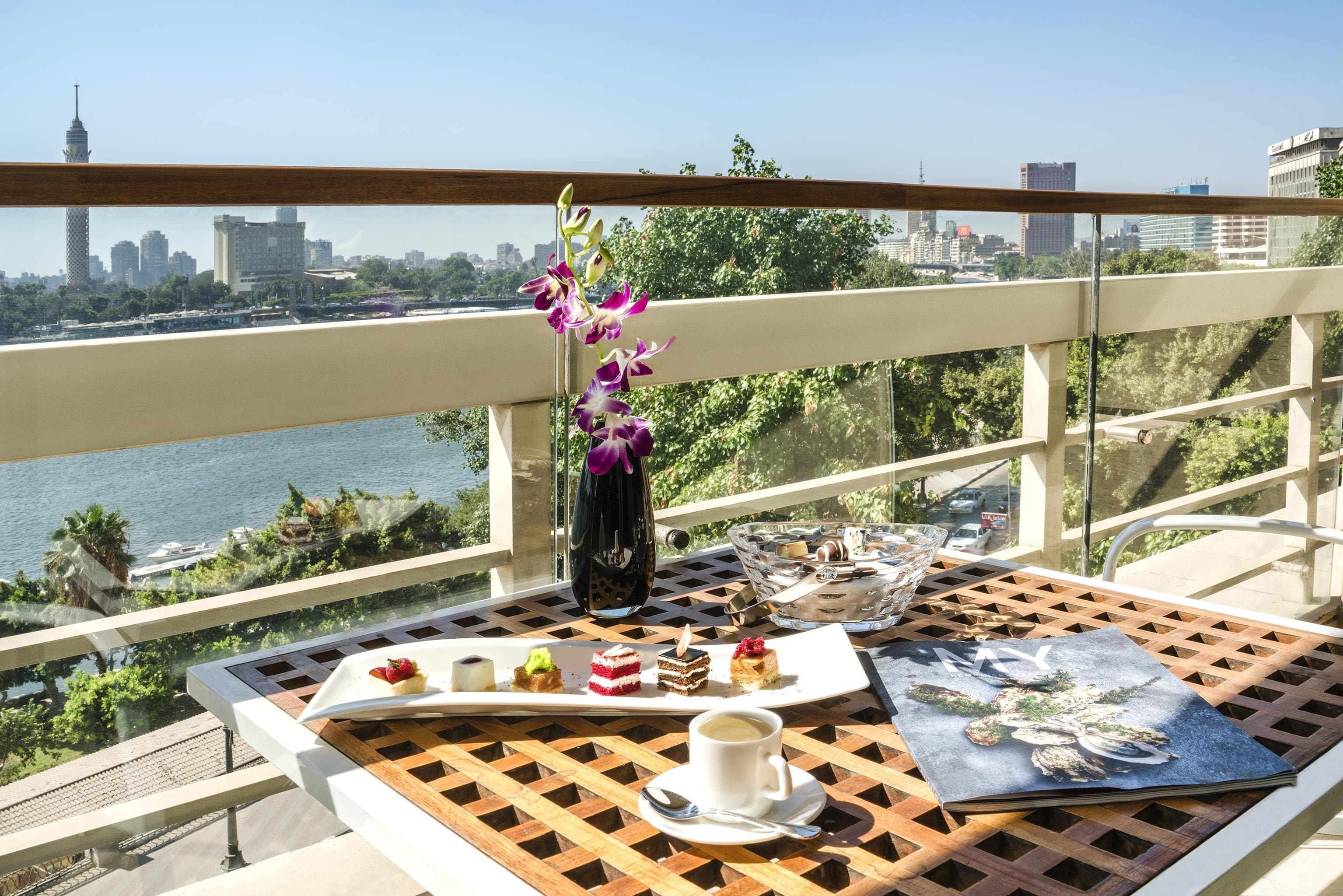 Un balcón con una mesa dispuesta para una comida, un jarrón con flores y vistas a un paisaje urbano.