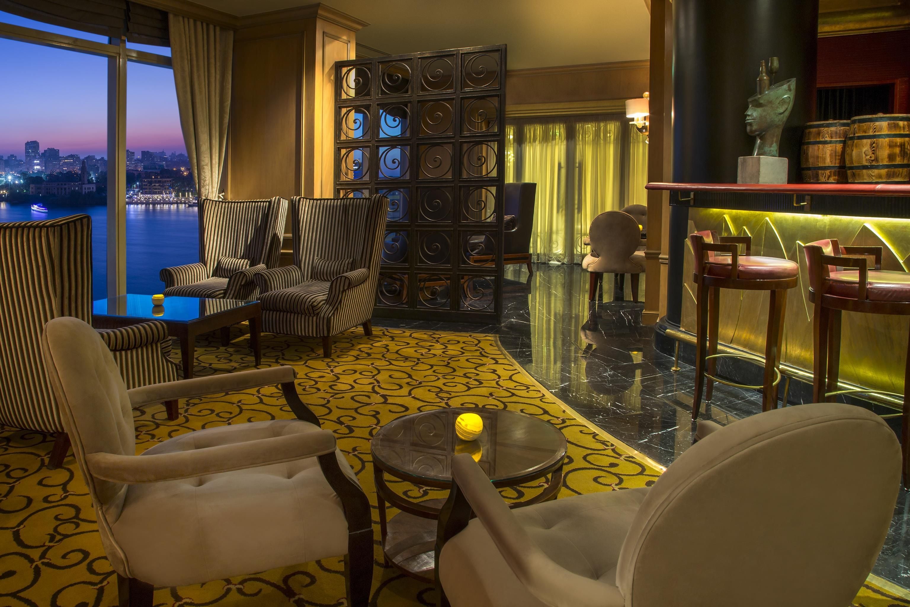 Habitación de hotel con vistas a un paisaje urbano al anochecer, con sillones a rayas, una pequeña mesa redonda y un separador decorativo de metal.