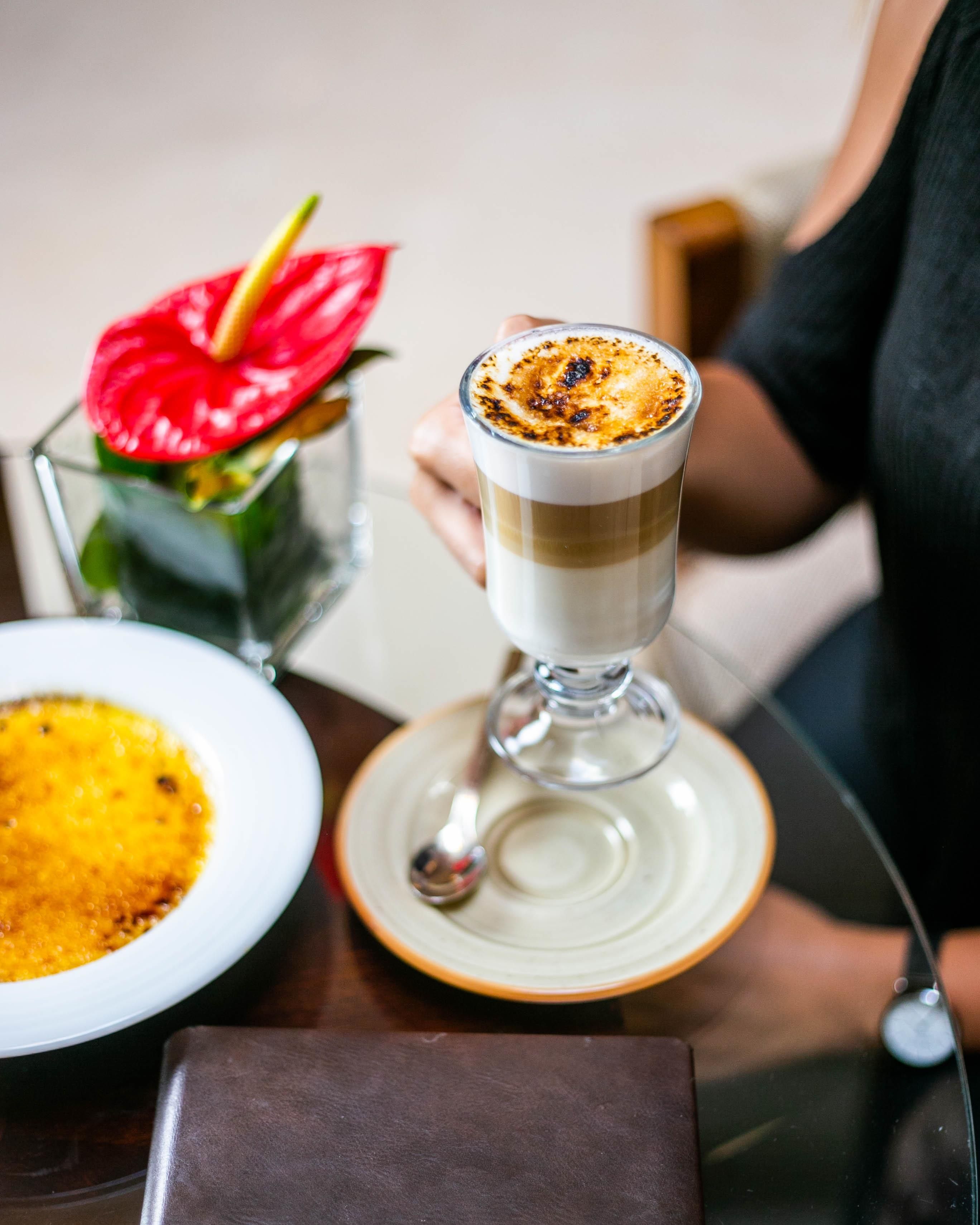 Un verre à dessert contenant une boisson en couches est posé sur une table, accompagné d’une assiette avec un dessert jaune et d’une fleur rouge dans un vase en verre.