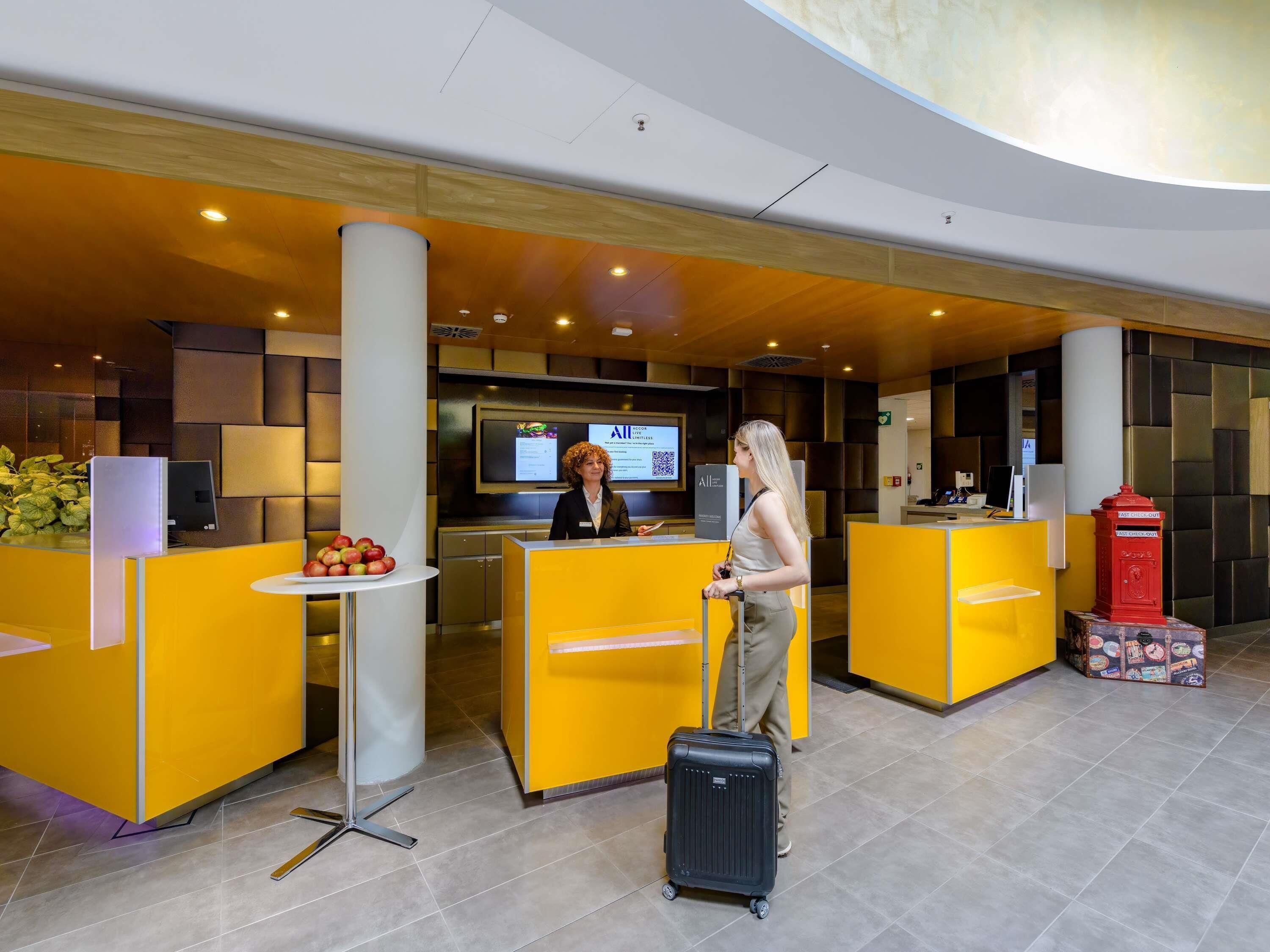 A hotel reception area with a modern design, featuring a reception desk, a digital screen, and a red fire alarm box.