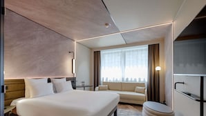 A modern hotel room with a large bed, a sofa, a small table, a TV, and a bathroom. - Novotel München Airport (Freising)