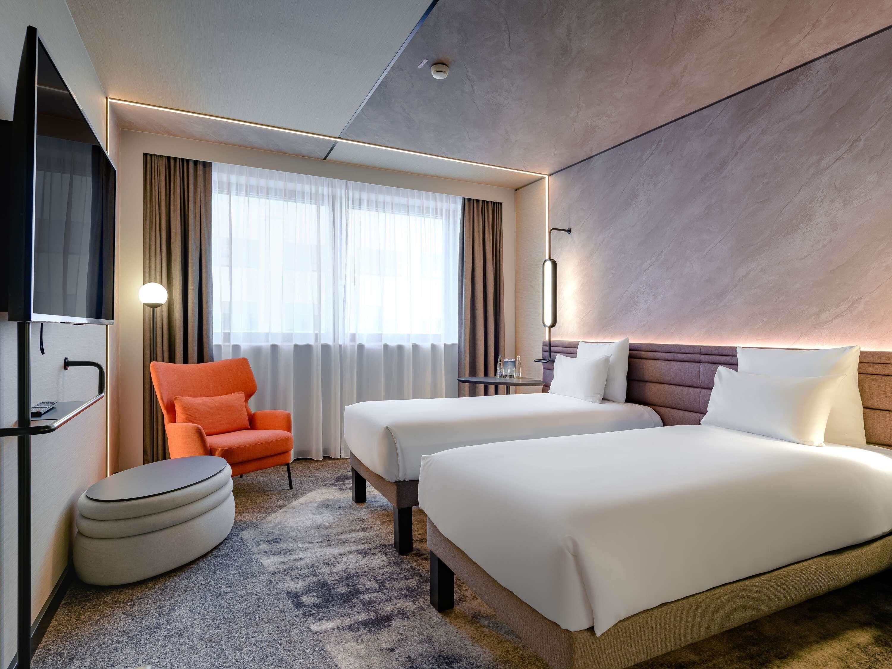 A hotel room with two beds, a TV, a chair, a small table, and a window with curtains.