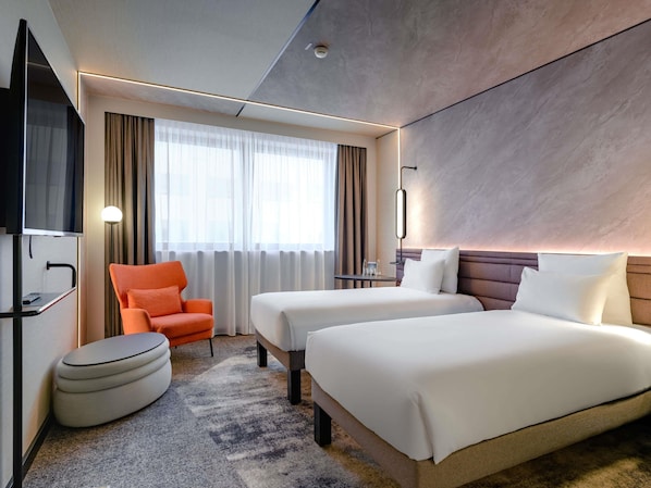 A hotel room with two beds, a TV, a chair, a small table, and a window with curtains.