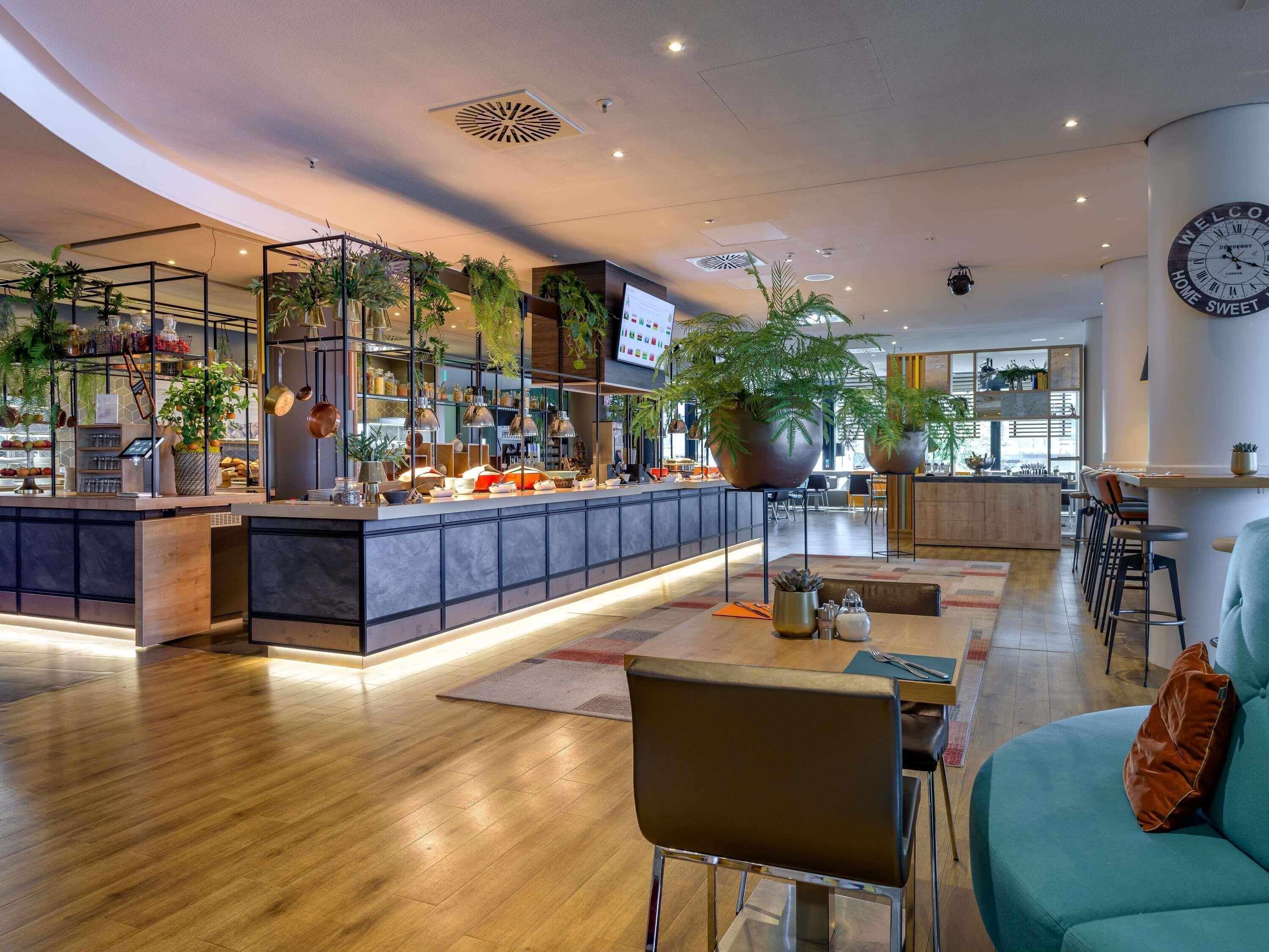 En moderne hotellobby med et centralt barområde, siddepladser og store plantekrukker.