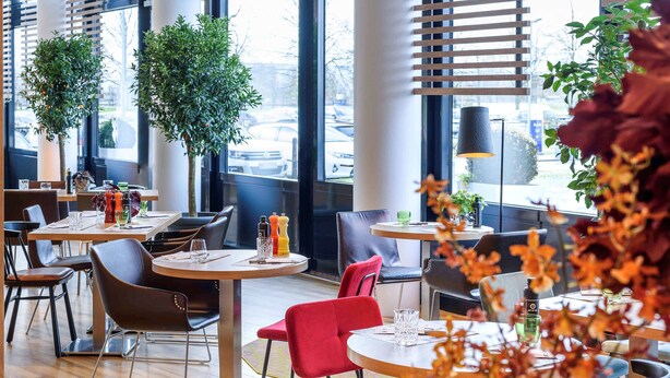 A modern restaurant interior with round tables, red and brown chairs, and potted plants. - Novotel München Airport (Freising)