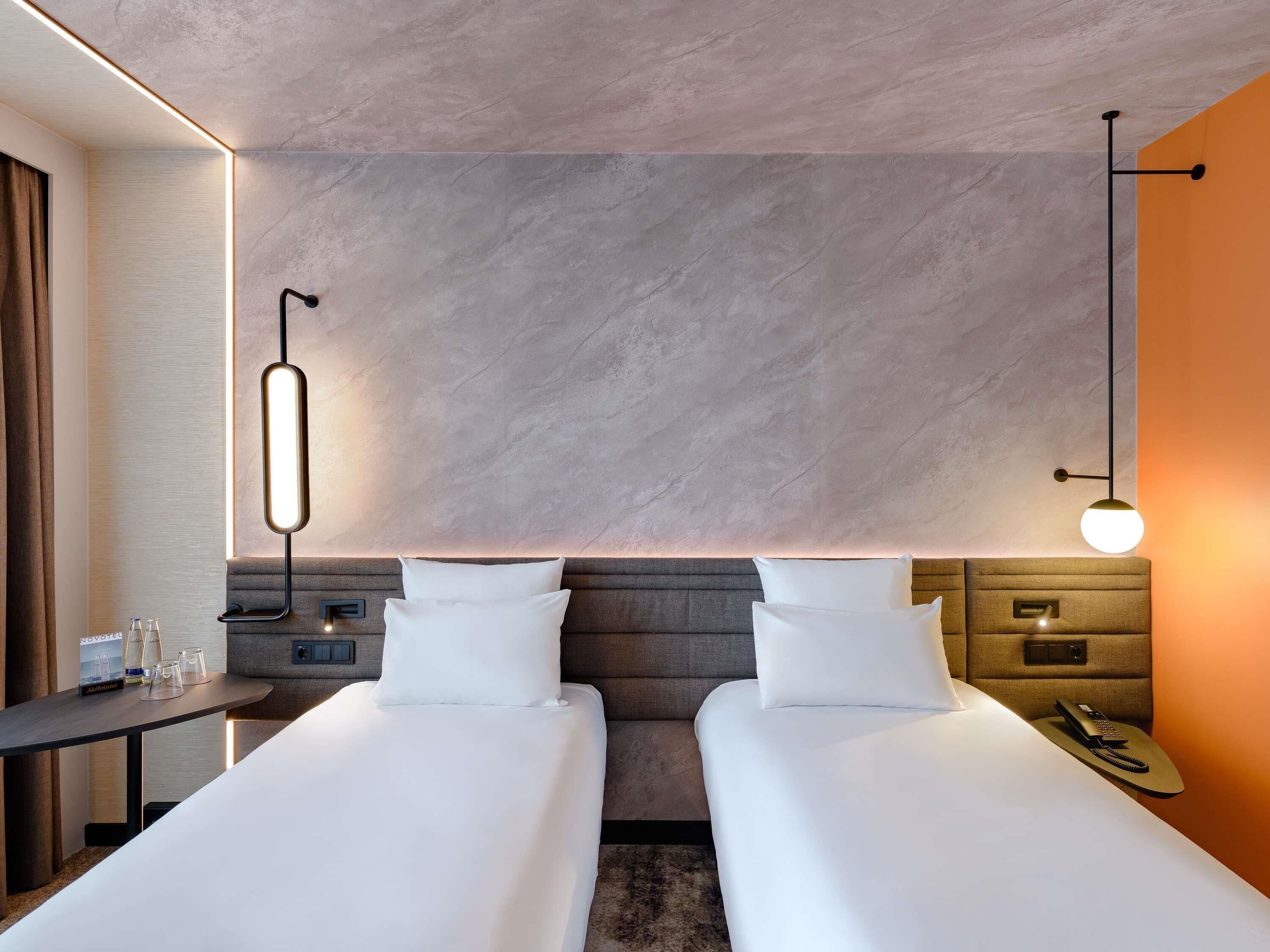 A hotel room with two beds, a desk with a lamp, a phone on the bedside table, and a wall-mounted light fixture.