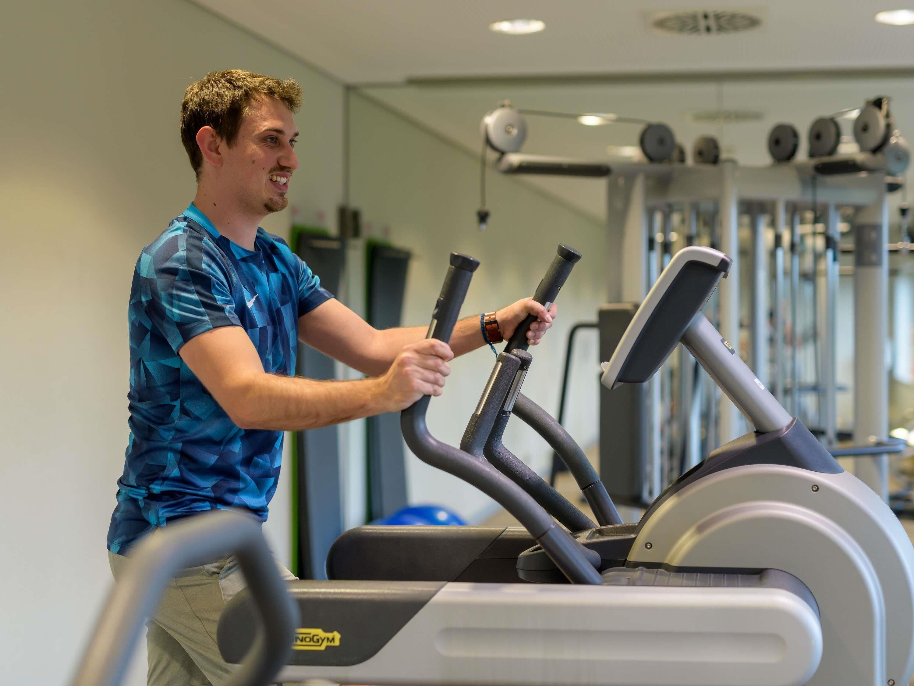 Een persoon gebruikt een elliptische trainer in een sportschool, met trainingsapparatuur op de achtergrond.
