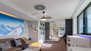 A modern living room with a sofa, coffee table, and a large mountain painting on the wall. - Novotel München Airport (Freising)