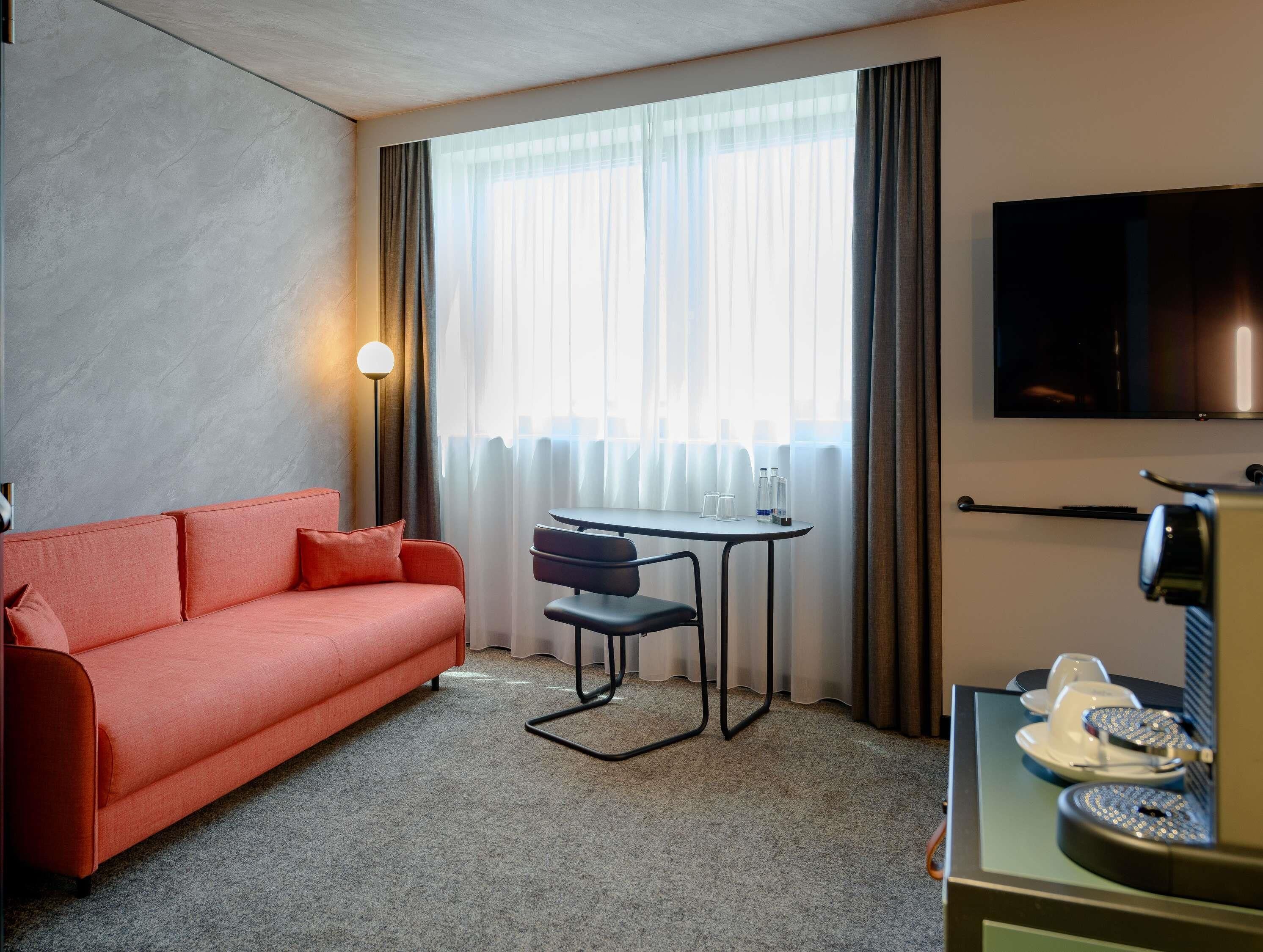 A hotel room with a sofa, a small table with chairs, a TV, and a minibar.