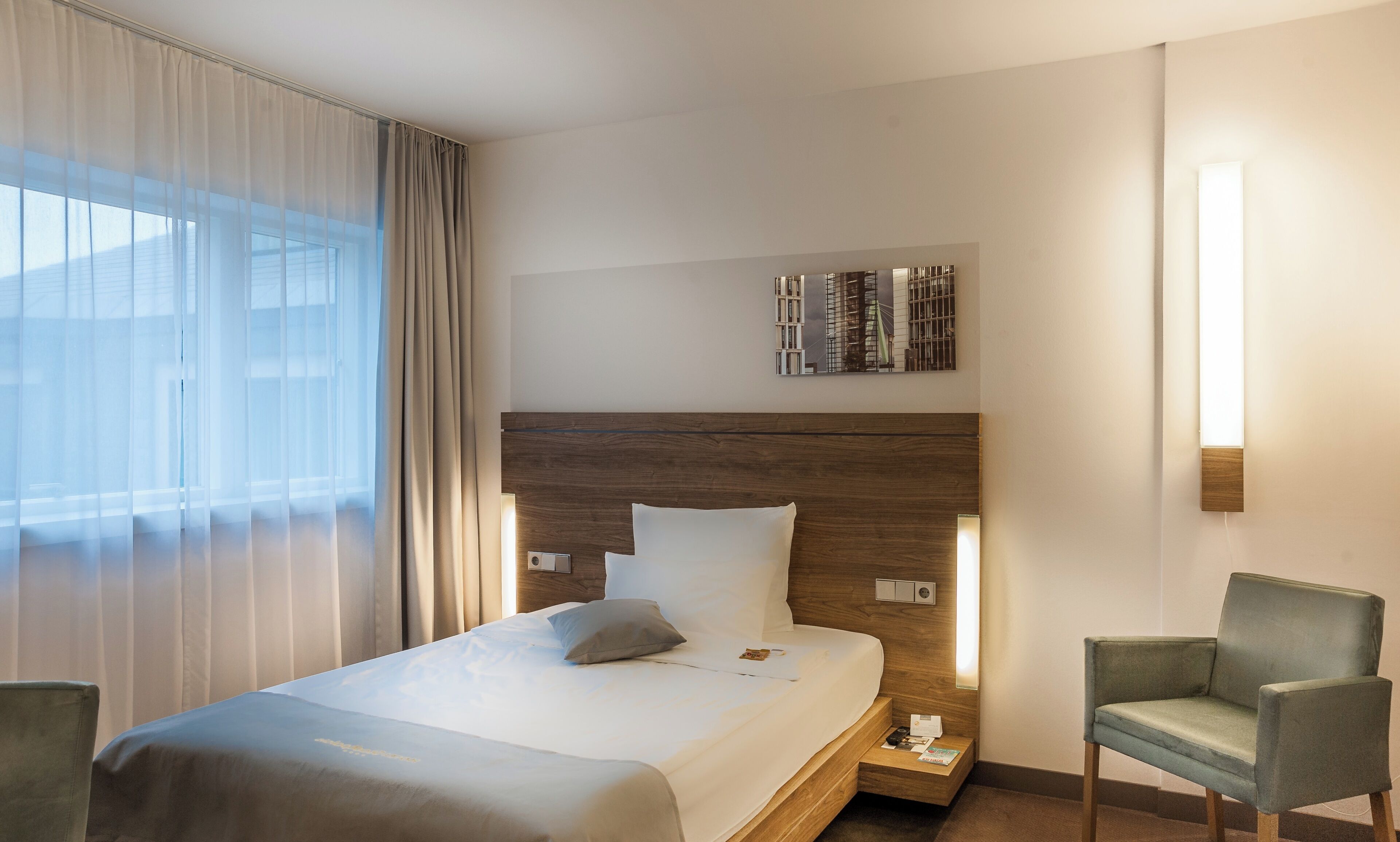 Una camera d'albergo con un letto, una sedia, un comodino e una lampada a parete.