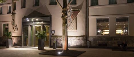 A hotel entrance with a glass door, a tree in the foreground, and a stone wall with a checkered pattern.