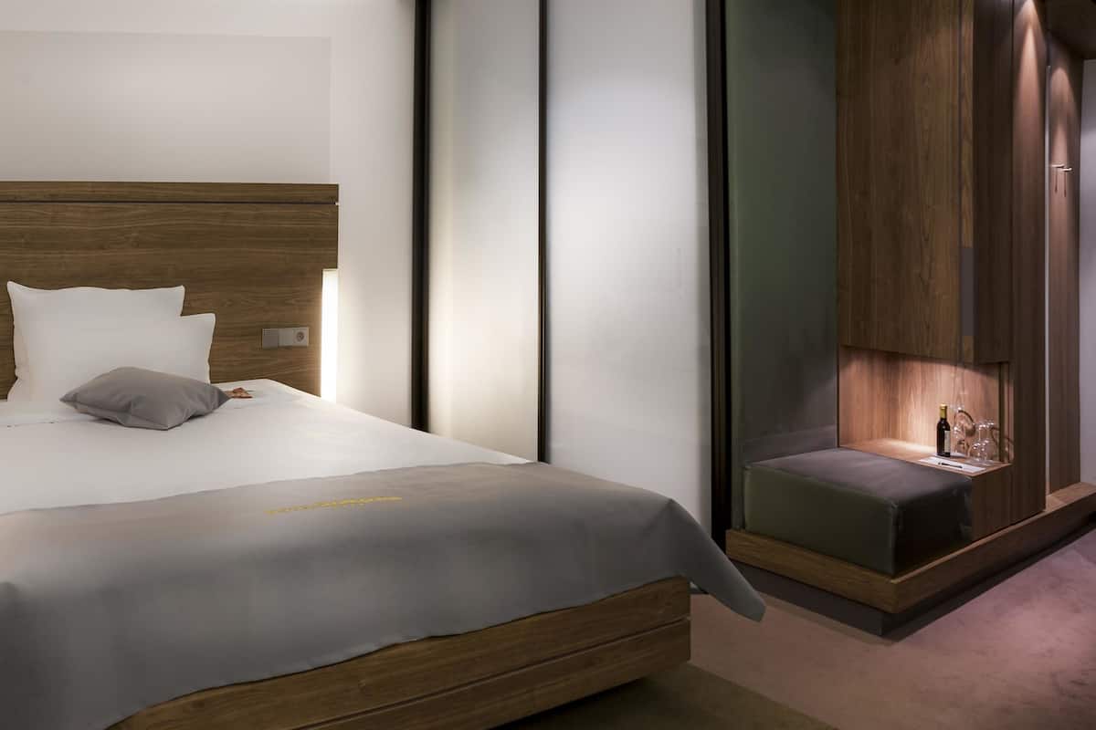 Quarto de hotel moderno com cabeceira de madeira, cama com roupa de cama branca, mesa de cabeceira com garrafa e copos, e um pequeno espaço de estar com um banco.