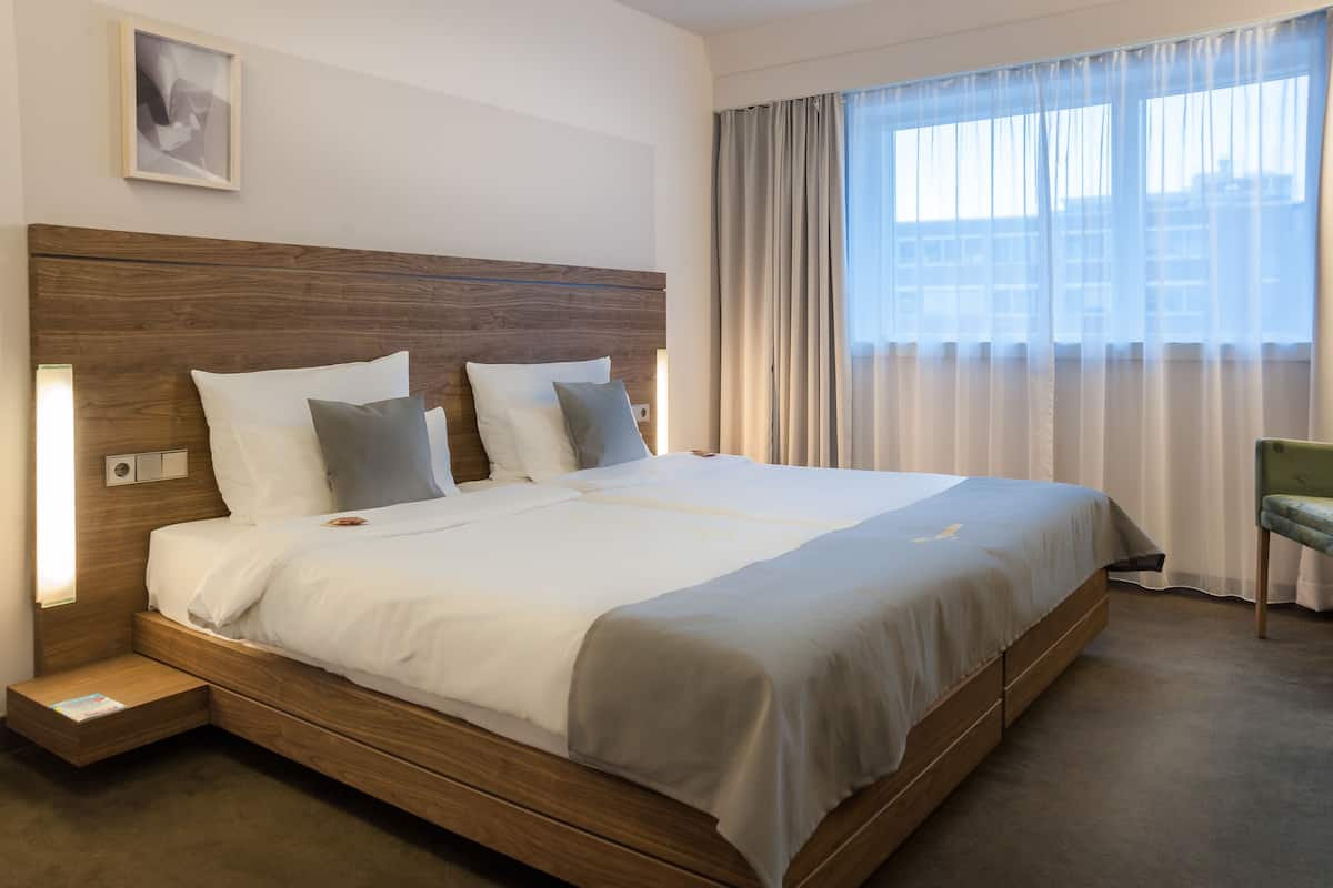 Quarto de hotel com uma cama grande, cabeceira de madeira, uma cadeira verde e uma janela com cortinas.