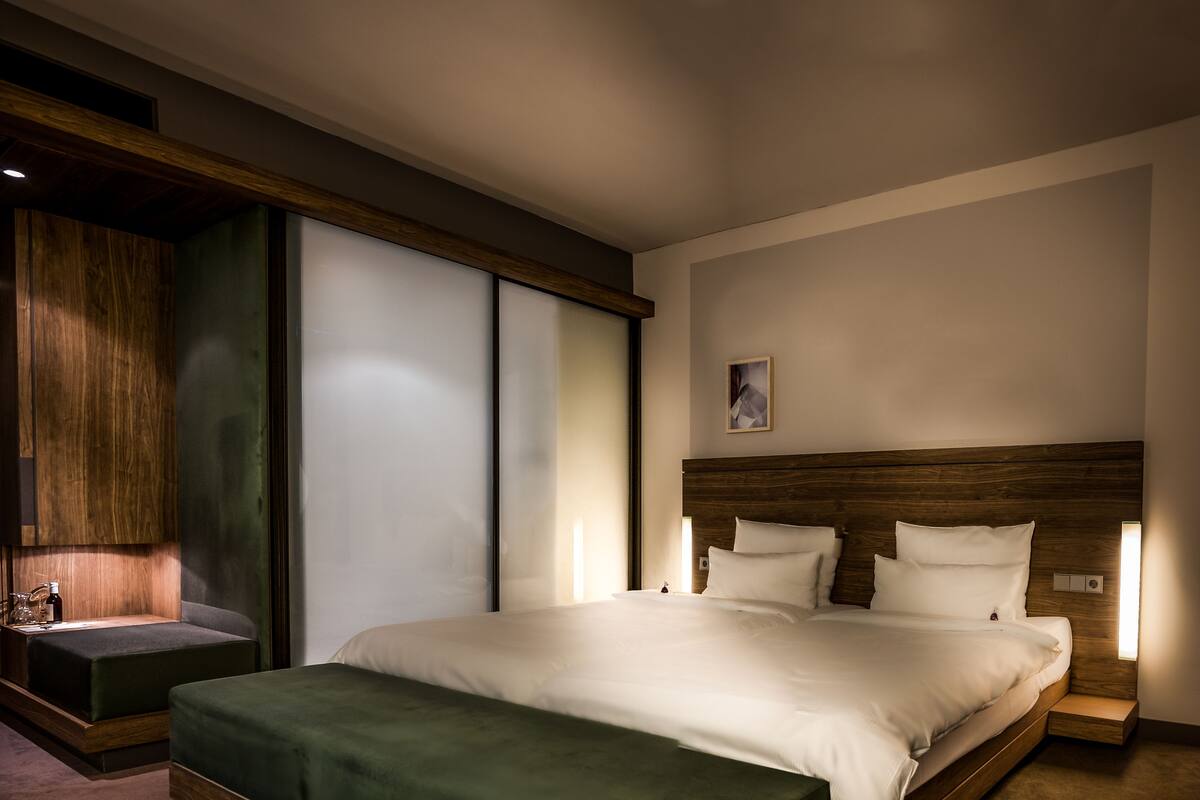 Quarto de hotel moderno com cama grande, cabeceira de madeira, área de estar com um banco e uma janela ampla.