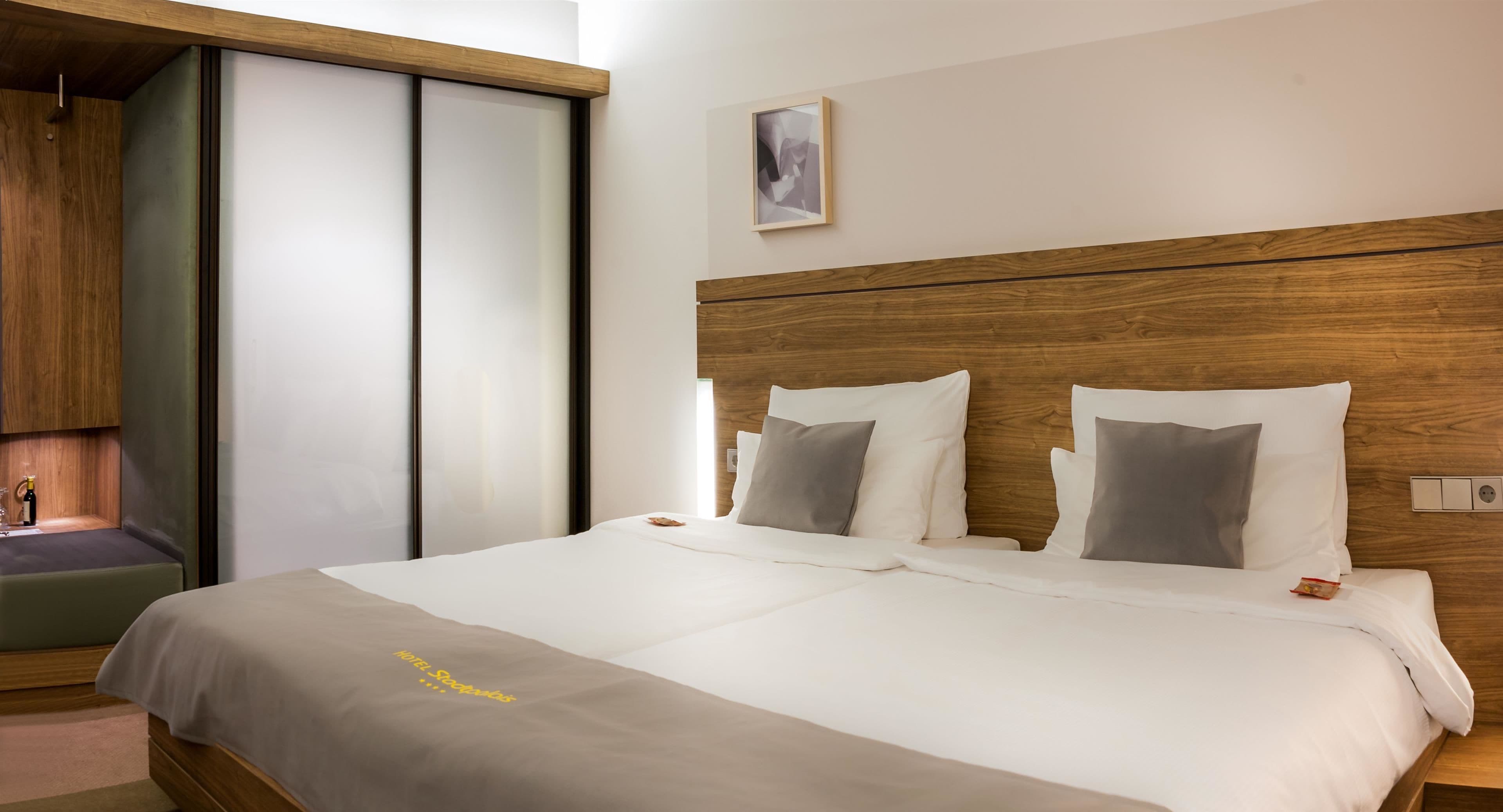 A hotel room with a wooden headboard, a bed with white and gray bedding, a bedside table, and a small window.