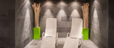 A spa area with two white lounge chairs, dark stone walls, and green planters with tall grasses.