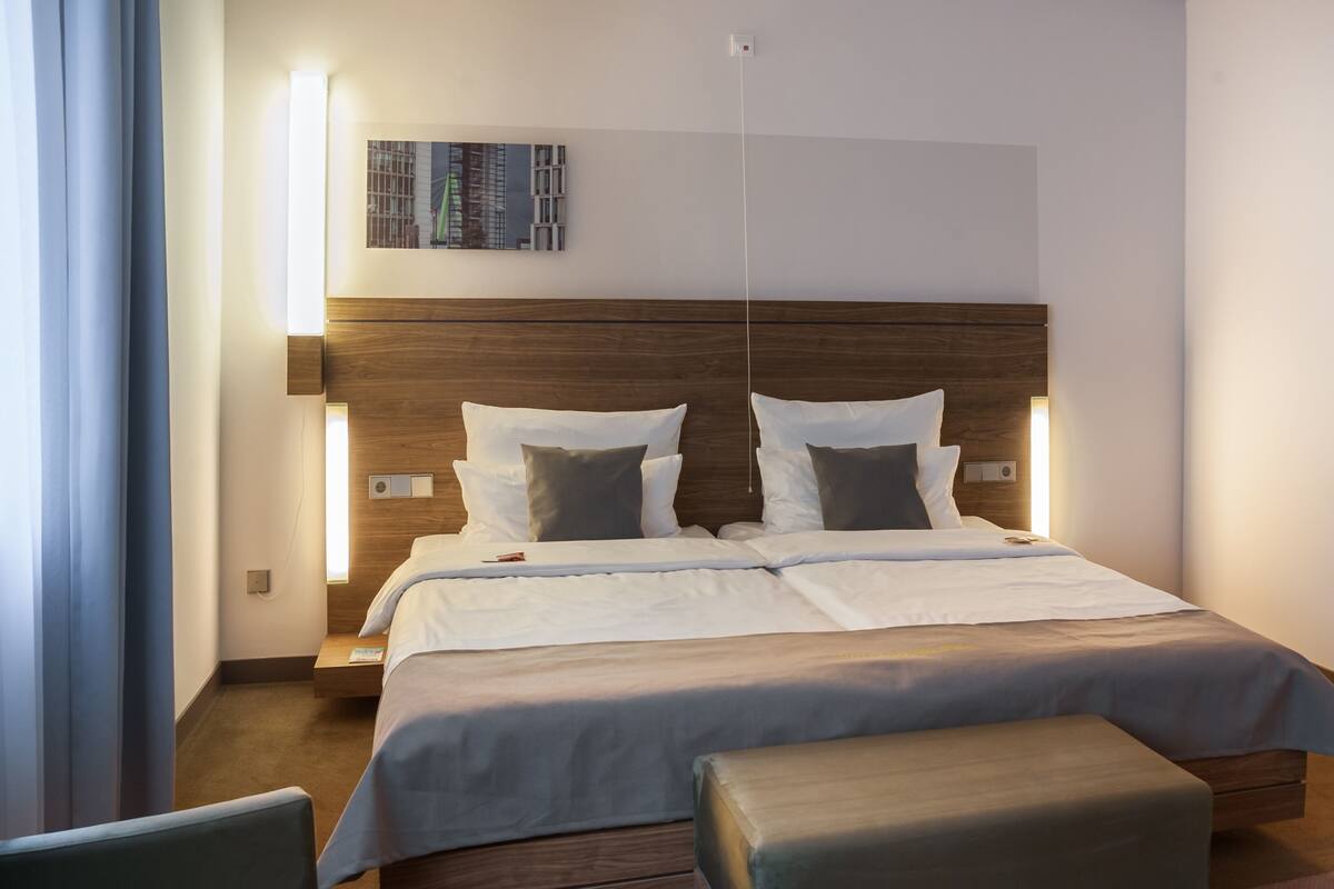 Quarto de hotel moderno com cabeceira de madeira, uma cama grande com roupa de cama branca, uma mesinha de cabeceira e um abajur pequeno.