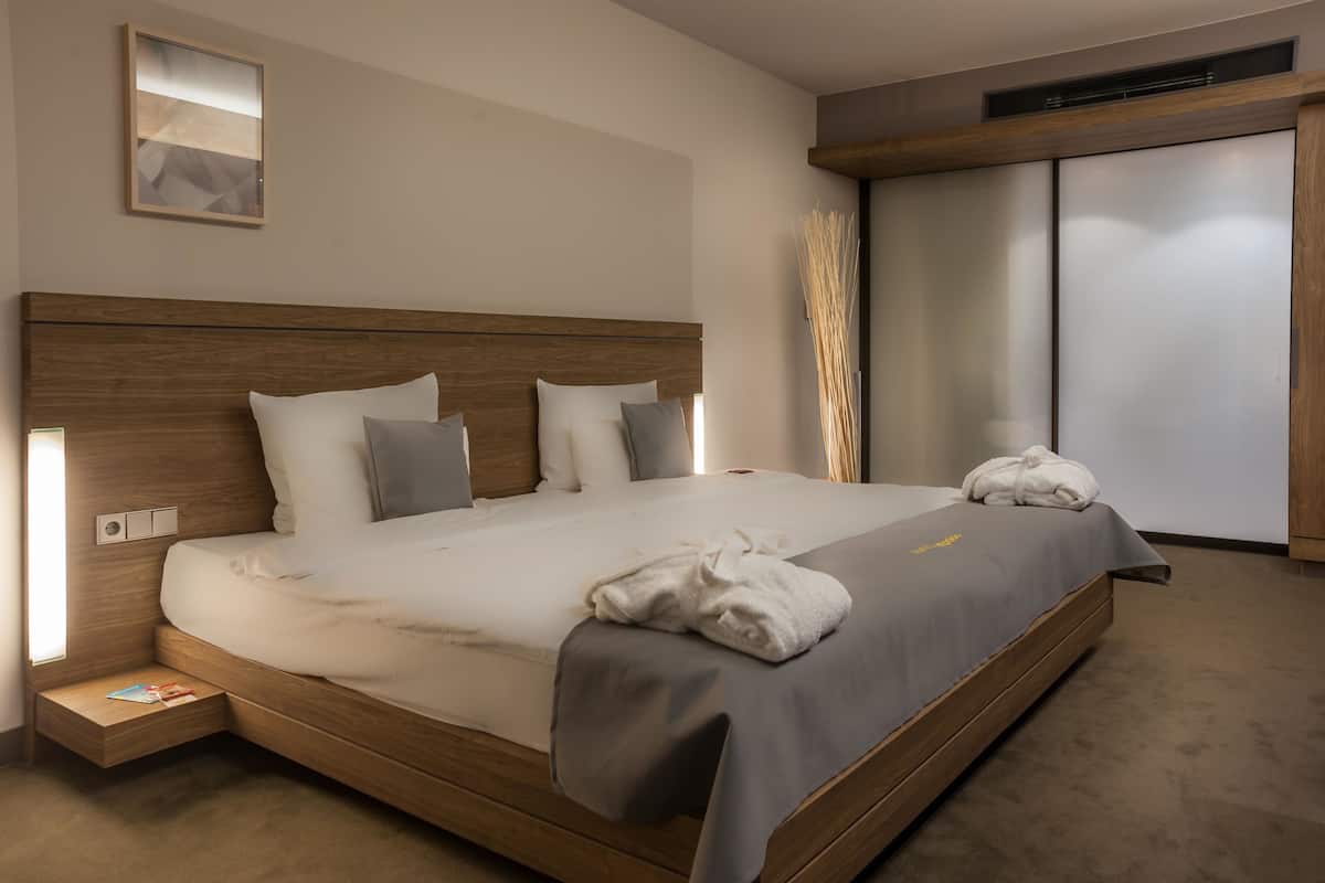 Quarto de hotel moderno com cabeceira de madeira, uma cama grande com roupa de cama cinzenta, mesa de cabeceira com abajur e guarda-roupa com portas de correr.
