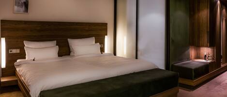 Quarto de hotel moderno com cama grande, cabeceira de madeira e um banco embutido com mesinha lateral.