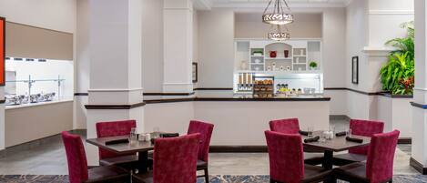 A hotel dining area with a central bar, red upholstered chairs, and a patterned carpet.