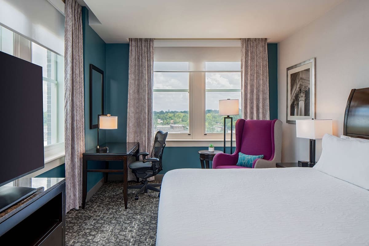 Ett hotellrum med en säng, ett skrivbord, en stol, en tv, en lampa, ett fönster med utsikt och en inramad tavla på väggen.