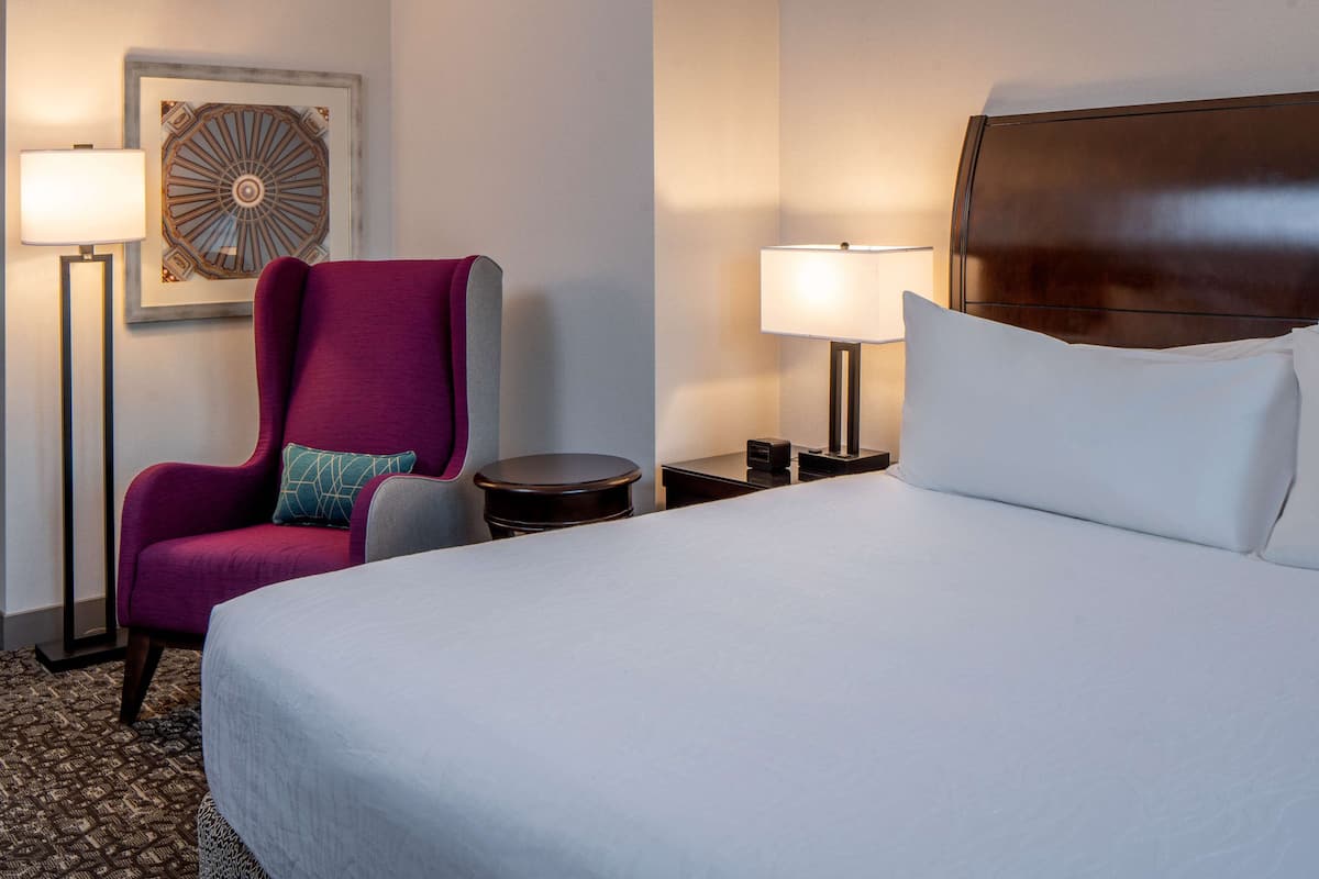 Ett hotellrum med en stor säng, en lila fåtölj, två sängbord med lampor och en dekorativ väggdekoration.