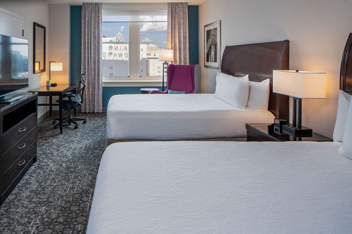 Ett hotellrum med två sängar, ett skrivbord, en stol, en TV och ett fönster med utsikt över staden.