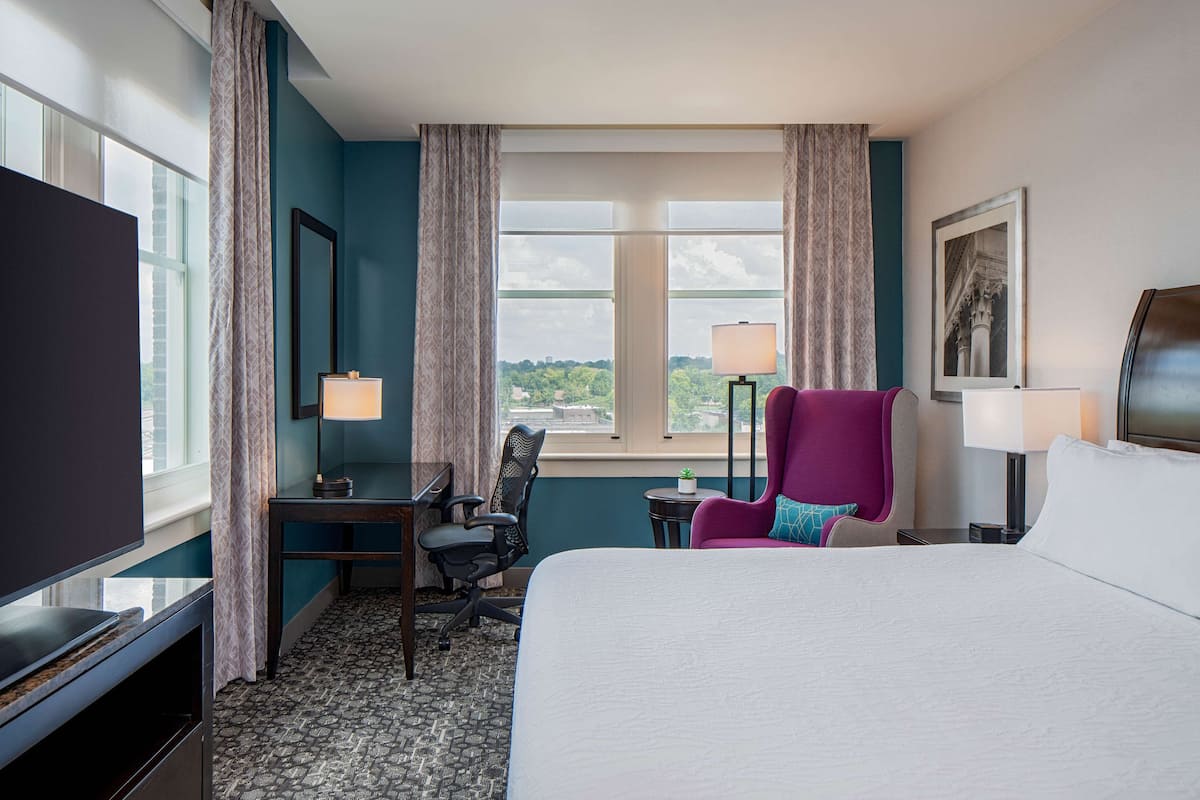 Ett hotellrum med en säng, ett skrivbord, en stol, en tv, ett fönster med gardiner och en lampa.
