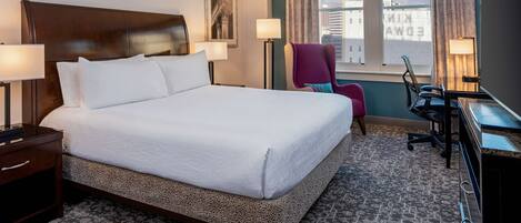 A hotel room with a large bed, two bedside tables, a desk with a chair, and a window with a view of buildings.